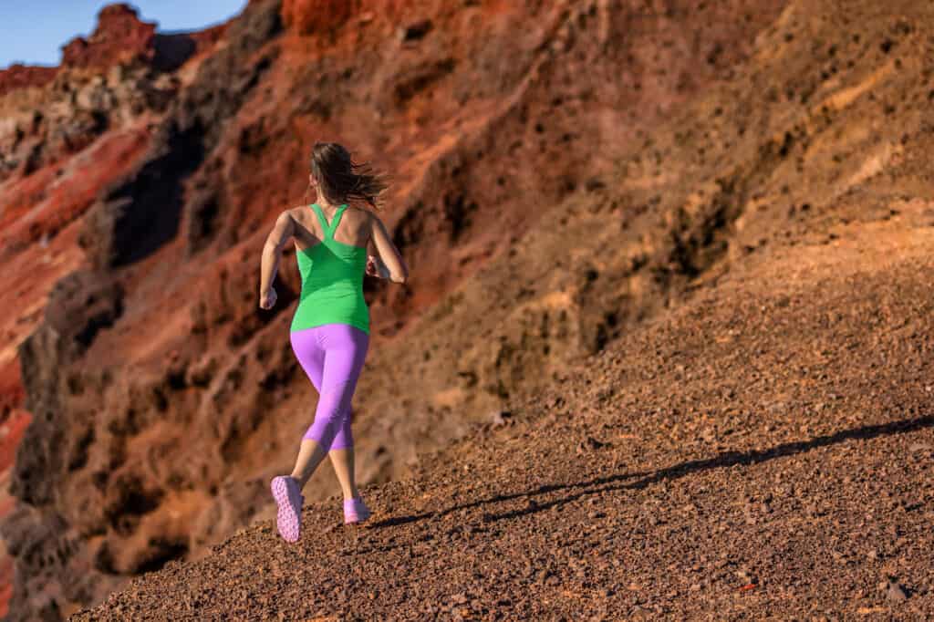 Trail runner woman ultra running uphill on volcano mountainside. Female athlete jogging in colorful activewear training hiit workout woman training outdoors.